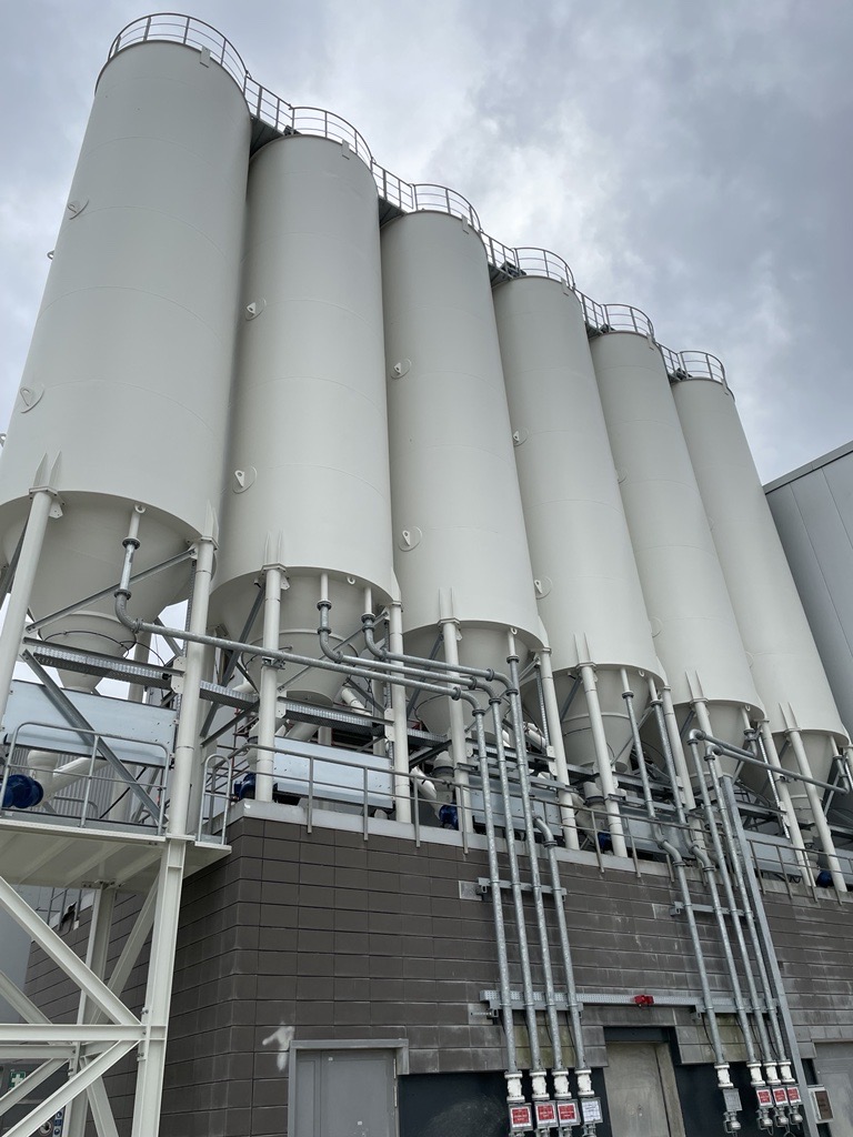 Ein Bild einer industriellen Betonmischanlage bei Tageslicht. Mehrere hohe, zylindrische Silos, die in einem sauberen Weiß gehalten sind, ragen in den bewölkten Himmel hinauf. Die Silos sind durch Metallrohre und -strukturen verbunden, die auf einer Betonplattform mit einer dunkelgrauen Wand im Hintergrund stehen. Unterhalb der Silos sind mehrere rote Notausschalter angebracht, und es gibt eine Metalltreppe auf der linken Seite, die zu einer Plattform und weiteren Bedienelementen führt.