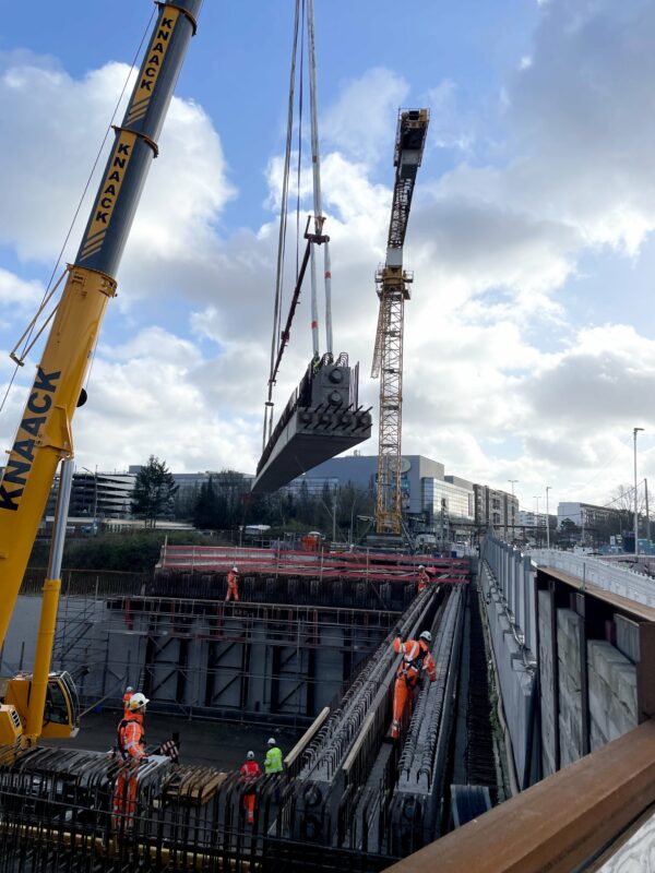 Arbeiter in Sicherheitskleidung auf einer Baustelle führen die Montage eines großen Betonfertigträgers mithilfe eines gelben Krans durch. Im Hintergrund ist ein teilweise im Bau befindlicher Tunnel zu sehen, mit Stahlarmierungen und weiteren Baumaschinen, darunter ein Turmdrehkran. Die Szenerie ist von einem bewölkten Himmel überdeckt, was auf die Komplexität und die Wichtigkeit der Sicherheitsvorkehrungen bei solchen Großprojekten hinweist.